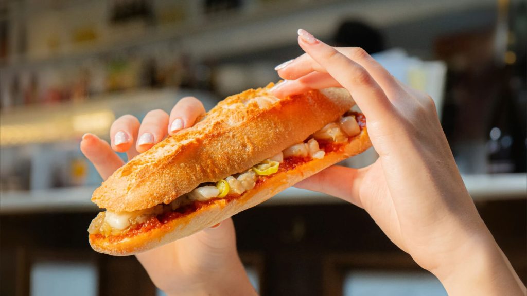 Mano sujetando el bocadillo de sobrasada, berenjena asada y queso de cabra. 