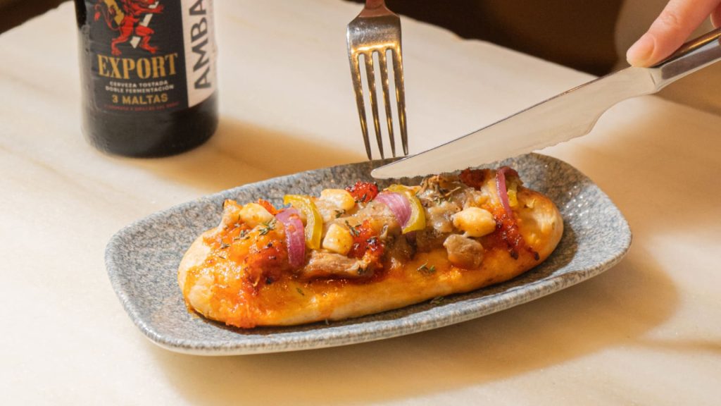 Coca con sobrasada, berenjena, queso de cabra, cebolla y pimiento encurtidos junto a un tercio de Ambar Export.