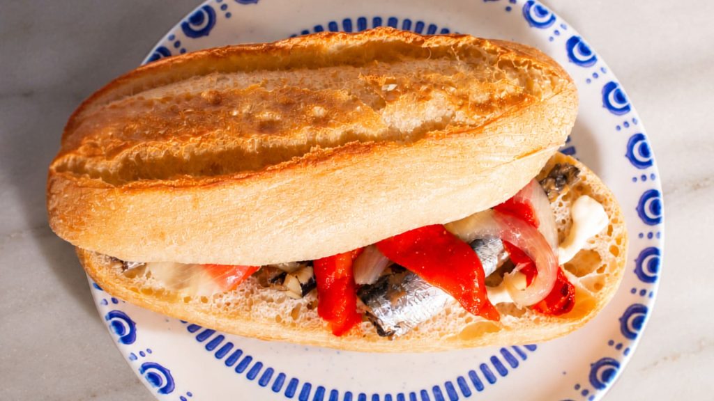 Bocadillo de sardinillas, pimientos asados, cebolla y mayonesa de ajo en plato de cerámica azul sobre mármol.