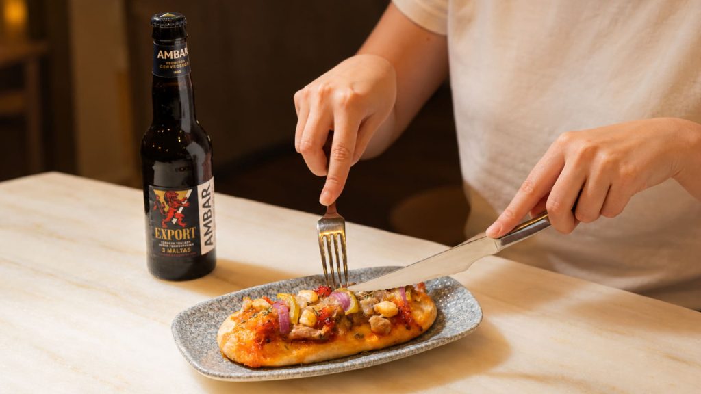 Comensal cortando la coca de Alenar de la Ruta de les Coques con un tercio de Ambar Export en una mesa de mármol. 
